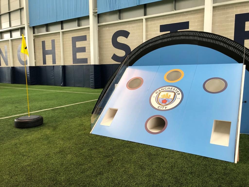 Curved sky-blue target board with scoring holes and a tyre arch, carrying the Manchester City crest